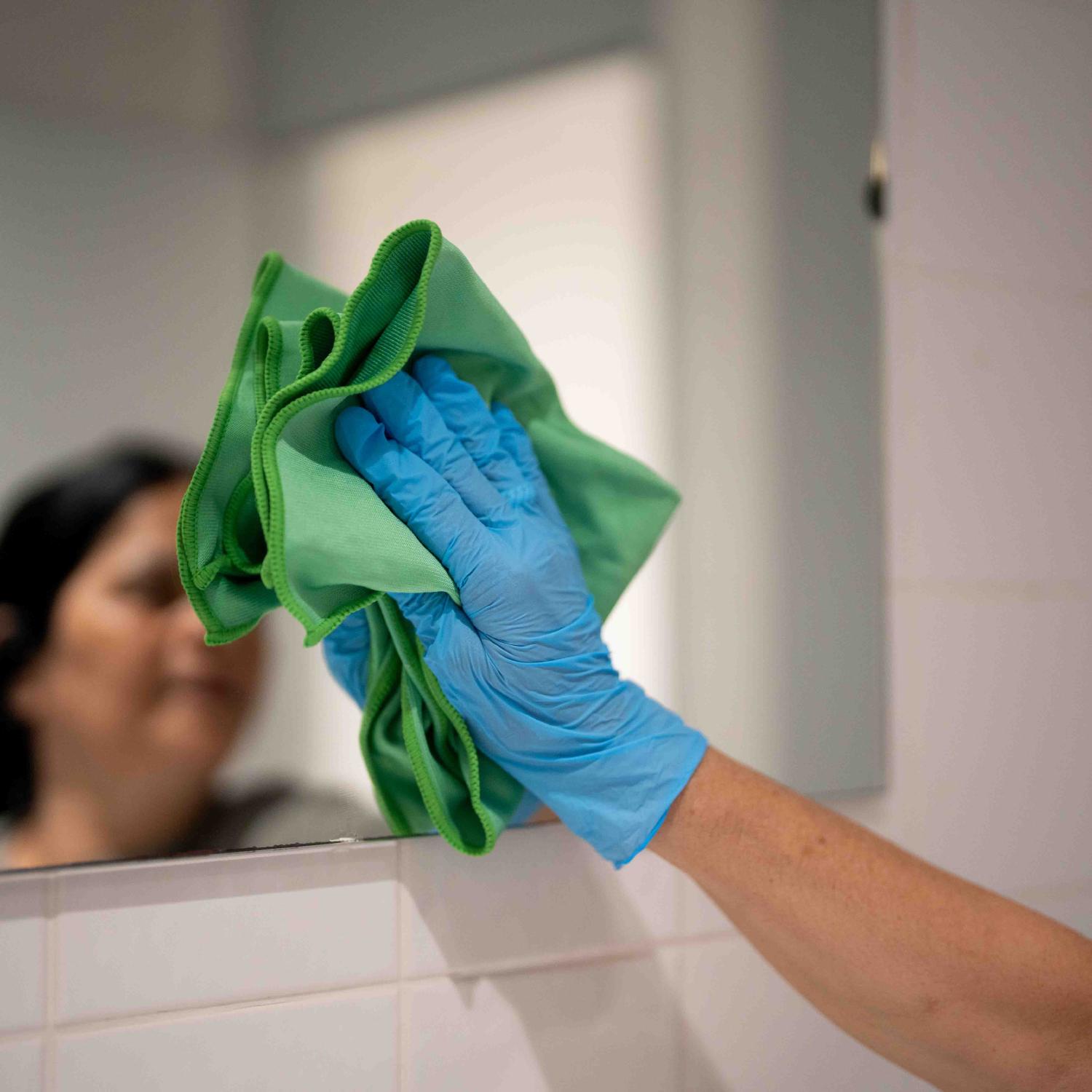 Close-up van handen met een groene microvezeldoek en blauwe handschoenen bij het schoonmaken van een spiegel
